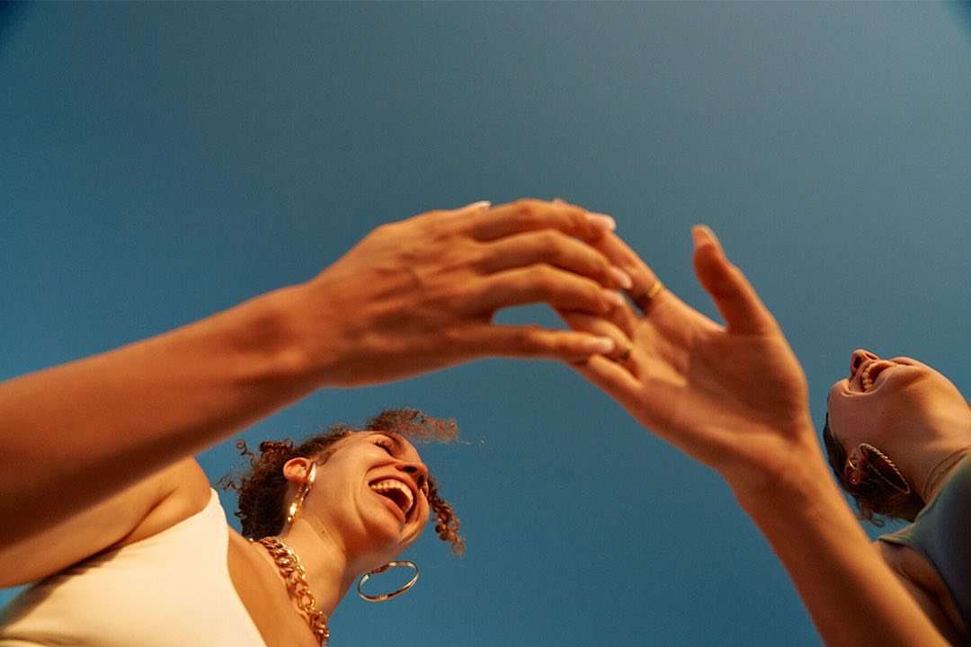Two women laugh in a low-angle shot against a bright blue sky, showcasing their faces, jewelry, and a sense of joy and connection.