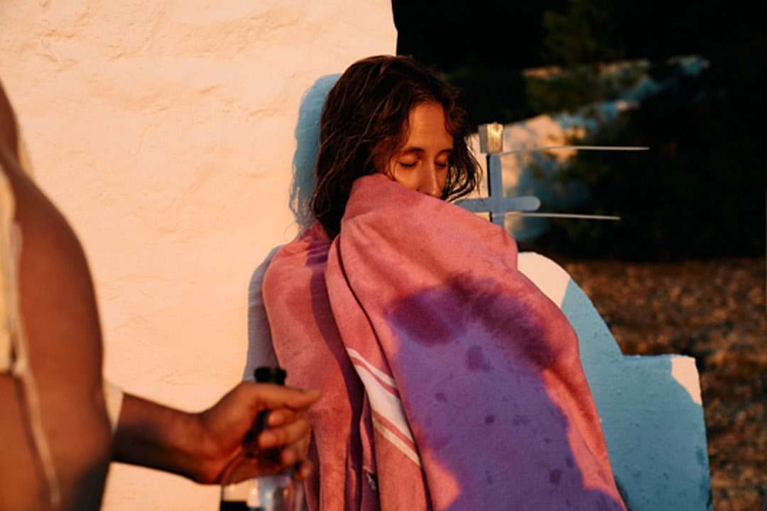 Woman wrapped in pink towel stands next to white wall with cross in outdoor setting during golden hour with hand reaching in.