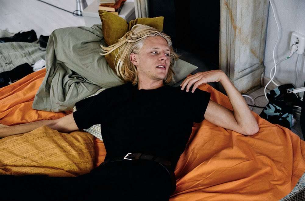 Blond man relaxing on a bed with an orange and yellow patterned blanket and pillows, wearing a black t-shirt in an indoor setting.