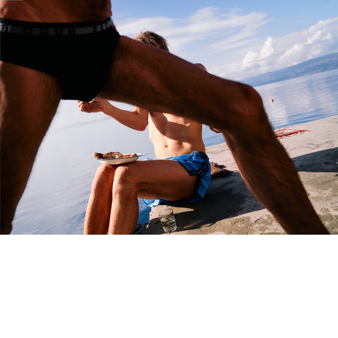 Two men on a concrete pier by the water, one eating pizza, blue sky, light clouds, legs framing view, lifestyle, outdoor setting.