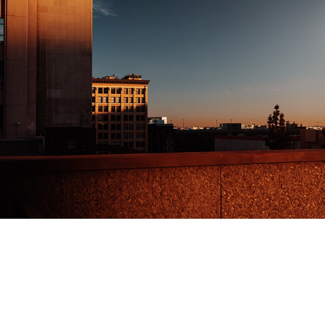 Urban skyline at sunset featuring golden-lit buildings against a clear sky, viewed from a rooftop edge. Architectural cityscape.