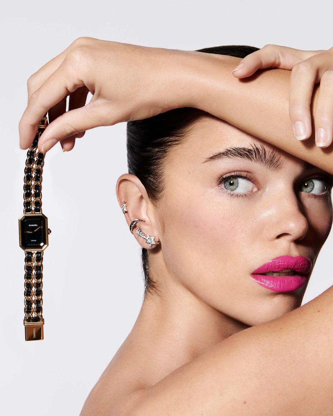 Close-up shot of an adult female model with pink lipstick holding a black and gold Chanel Première watch against a white background.