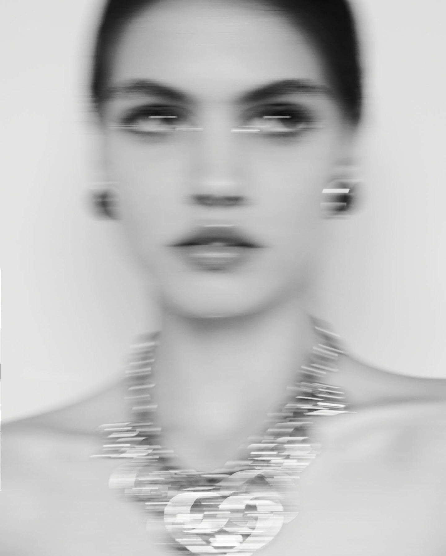 Monochromatic blurred portrait of an adult female wearing jewelry, including a necklace and earrings, against a light background.