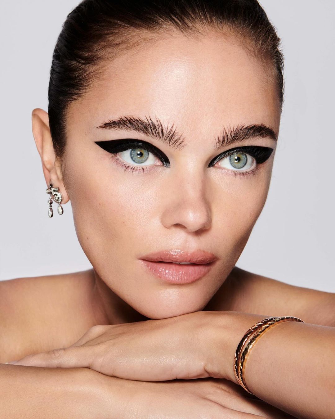 Close-up studio shot of a woman with graphic black winged eyeliner, bracelet jewelry, and bare shoulders against a neutral backdrop.