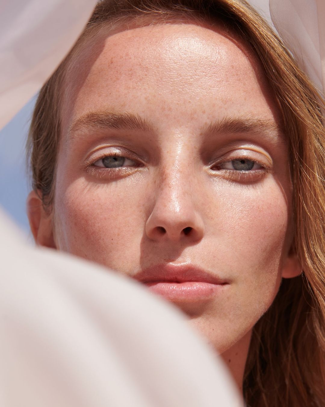 Close-up portrait of a fair-skinned woman with red hair and blue eyes, partially veiled in sheer white fabric, under bright, natural lighting.