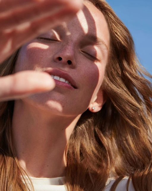 Portrait of fair-skinned woman shielding her eyes from the sun, eyes closed, with natural brown hair and a blue sky backdrop.