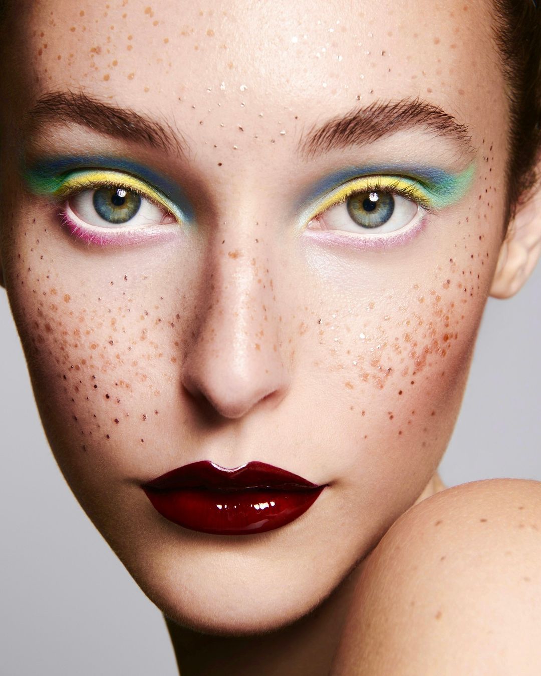 Close-up portrait of a woman with freckles wearing colorful eyeshadow and dark red lipstick against a gray background.