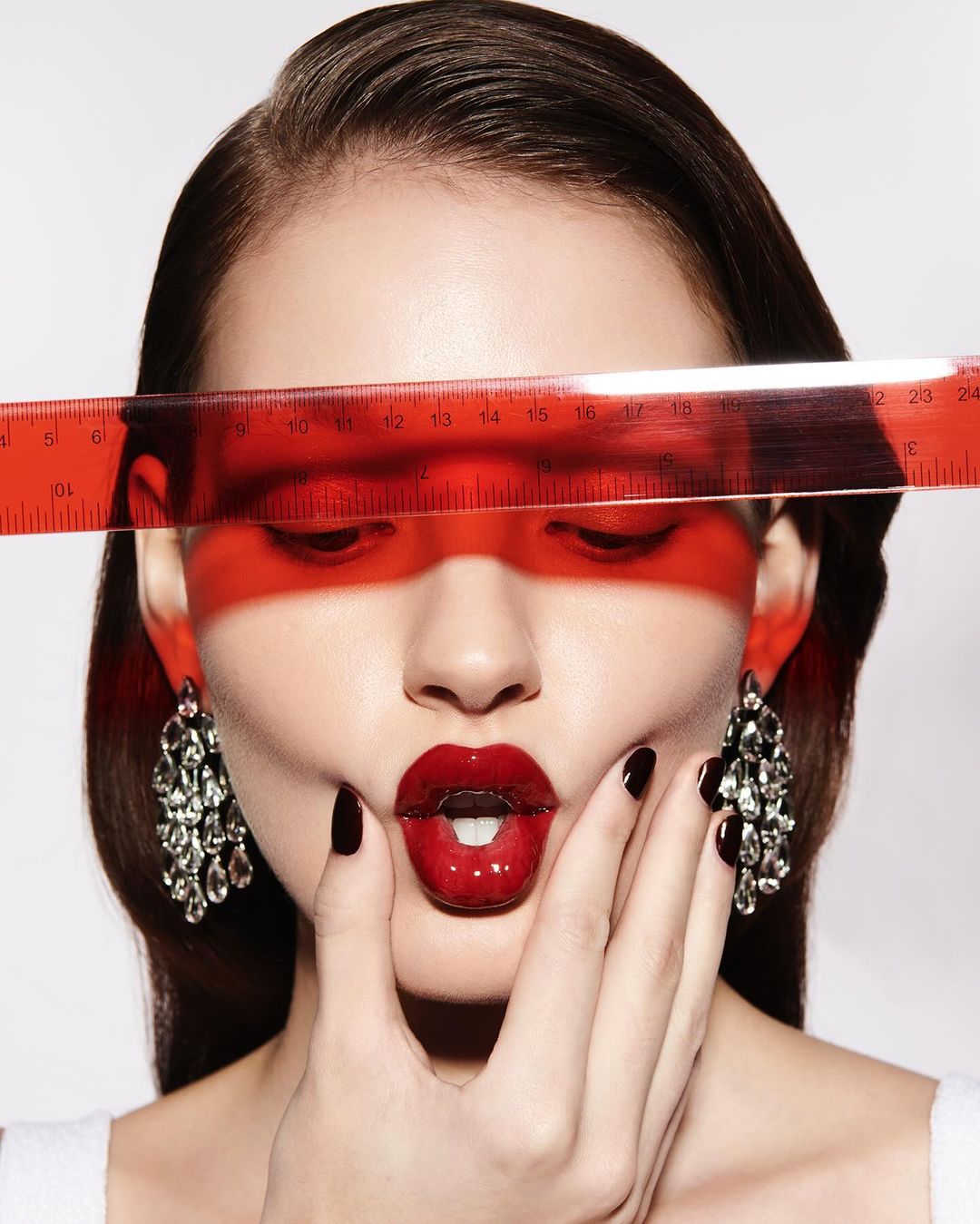 Close-up of a brunette woman with dramatic red lipstick, chandelier earrings, a ruler across her eyes, and manicured hands near her face.