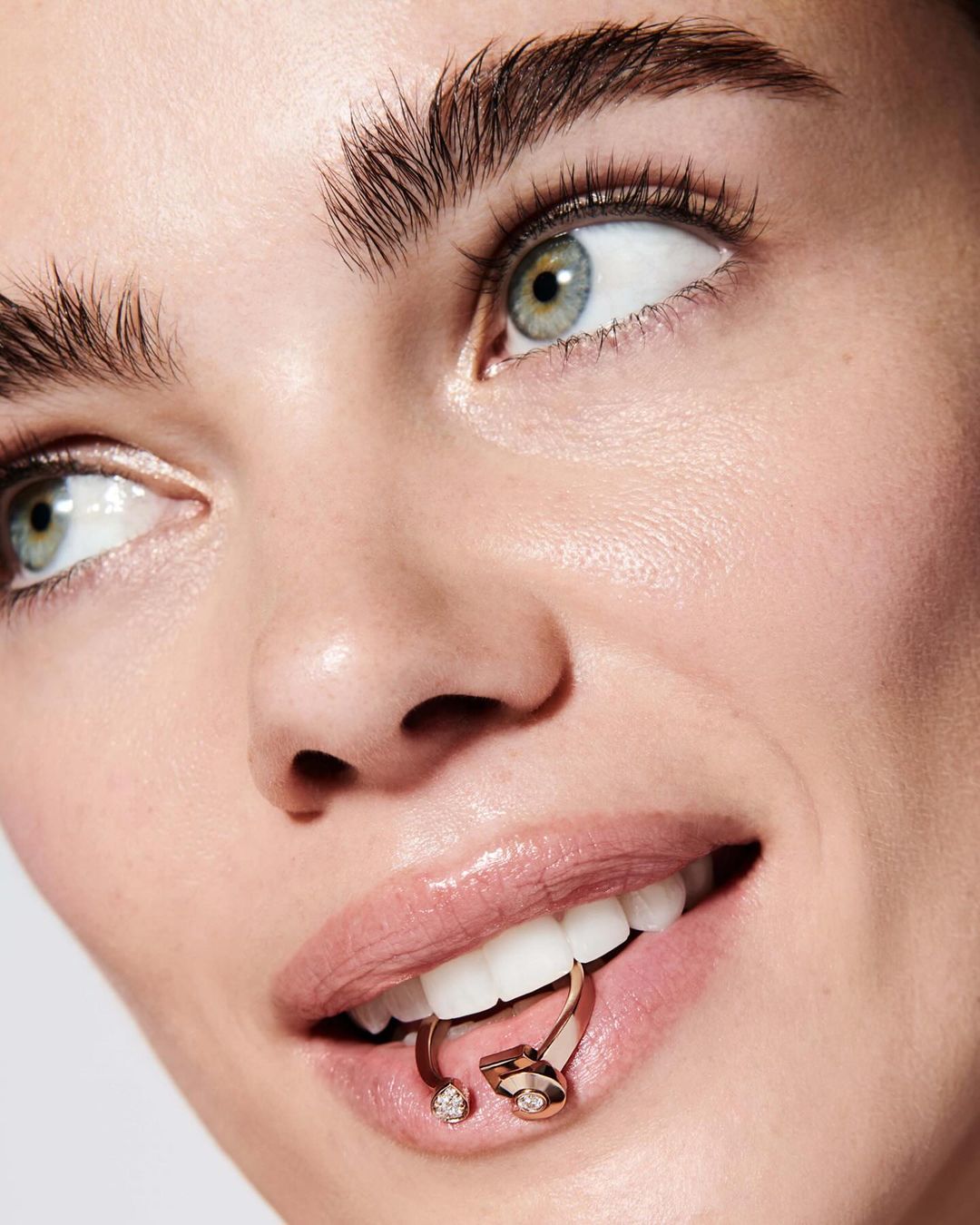Close-up of a woman's face with light skin, arched eyebrows, and a gold metal ring piercing held in her mouth between her front teeth.