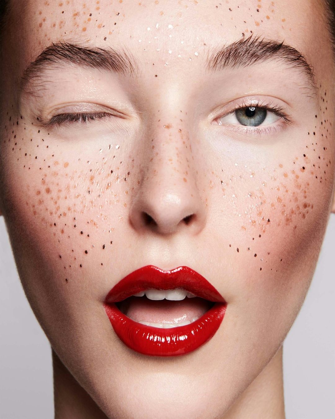 Close-up studio shot of a young adult female model with fair skin, freckles, one eye closed, and glossy red lipstick on light grey background.