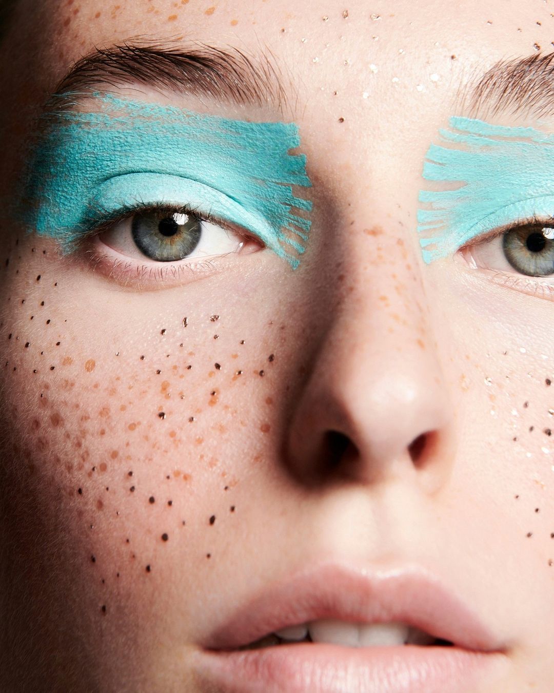 Close-up of a woman's face with freckles and vibrant turquoise makeup applied to her eyebrows, enhancing her light-colored eyes.