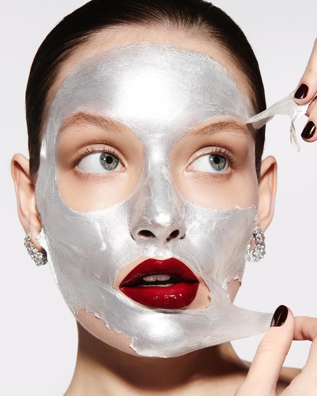 Woman removes silver facial mask; beauty shot shows cosmetics applied with red lipstick, with fair skin and decorative earrings.
