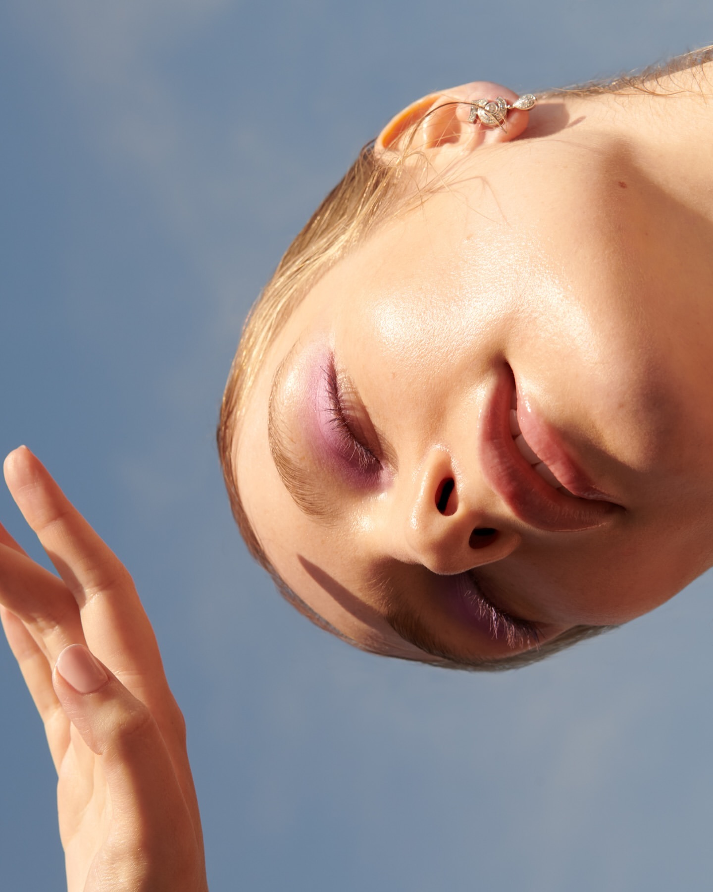 Eye-level portrait of a woman with closed eyes and lilac makeup against a serene blue sky, highlighting her face and an open hand.