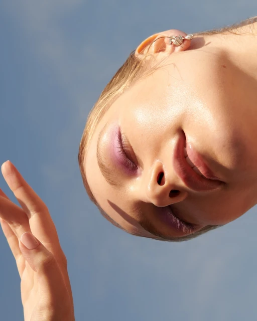 Eye-level portrait of a woman with closed eyes and lilac makeup against a serene blue sky, highlighting her face and an open hand.