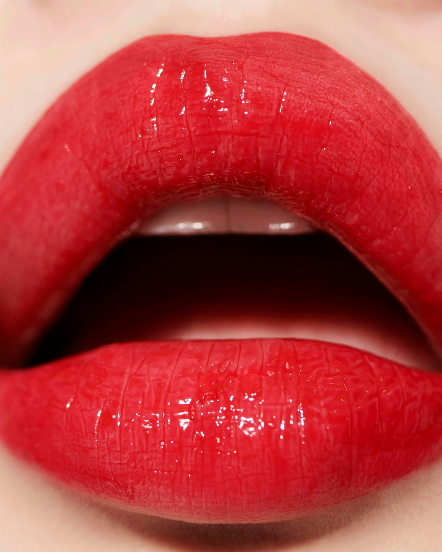 Close-up of a woman's red lips with glossy lipstick, showing teeth in an indoor setting. Beauty shot focuses on vivid color and detail.