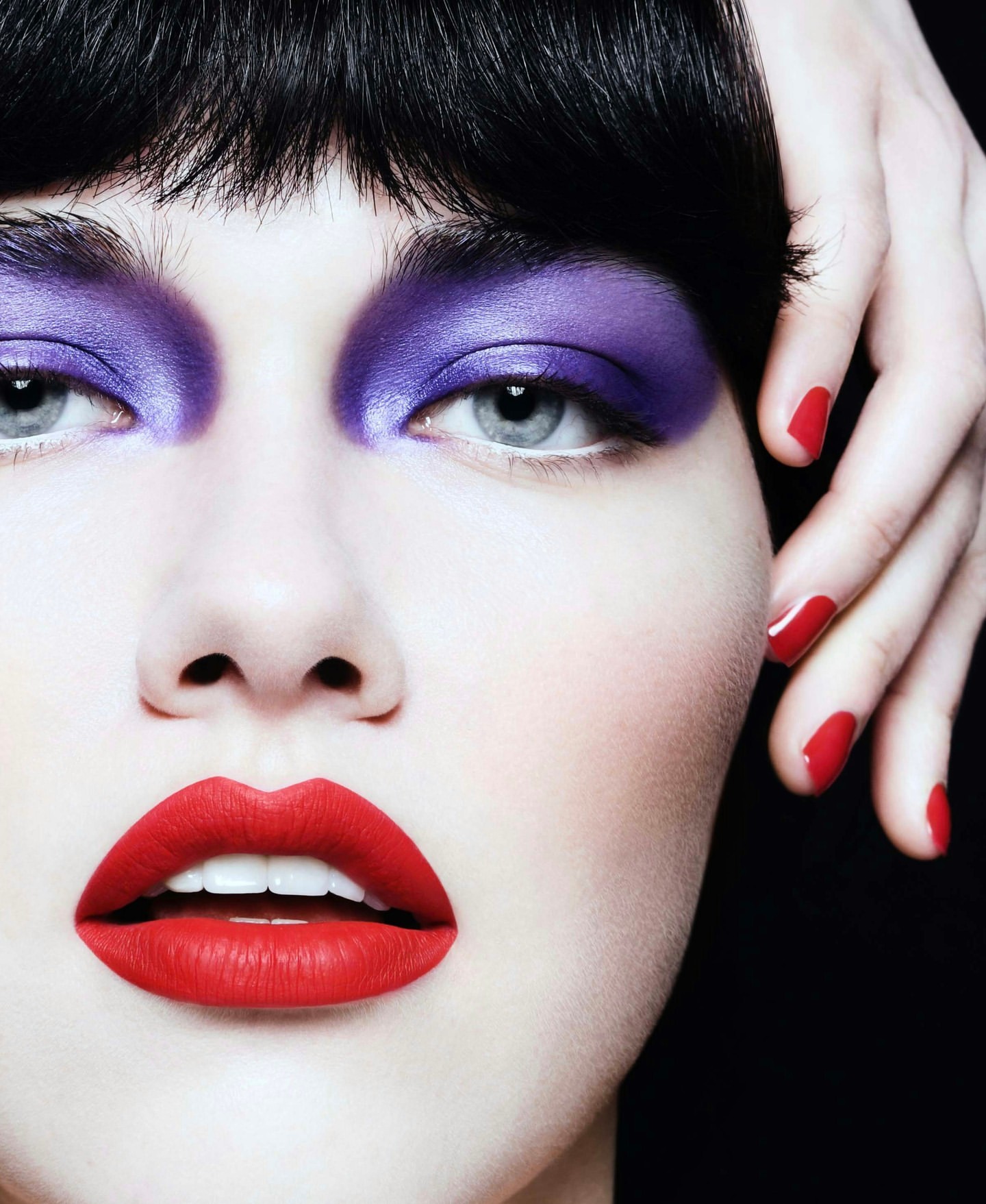 Close-up studio shot of a woman with bold purple eyeshadow, red lipstick, short dark hair, and manicured red nails against a dark background.