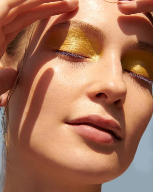 Close-up of a fair-skinned woman with fingers framing her face, eyes closed, wearing golden eyeshadow and blue mascara, in soft sunlight.