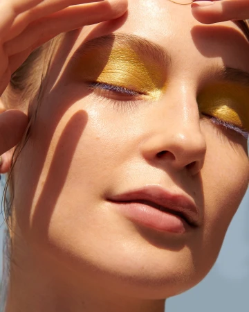 Close-up of a fair-skinned woman with fingers framing her face, eyes closed, wearing golden eyeshadow and blue mascara, in soft sunlight.