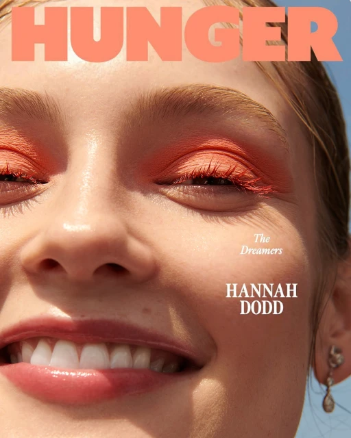 Close-up of a woman's face with coral makeup, including eyeshadow, and glossy lips, against a blue sky background, from Hunger Magazine.