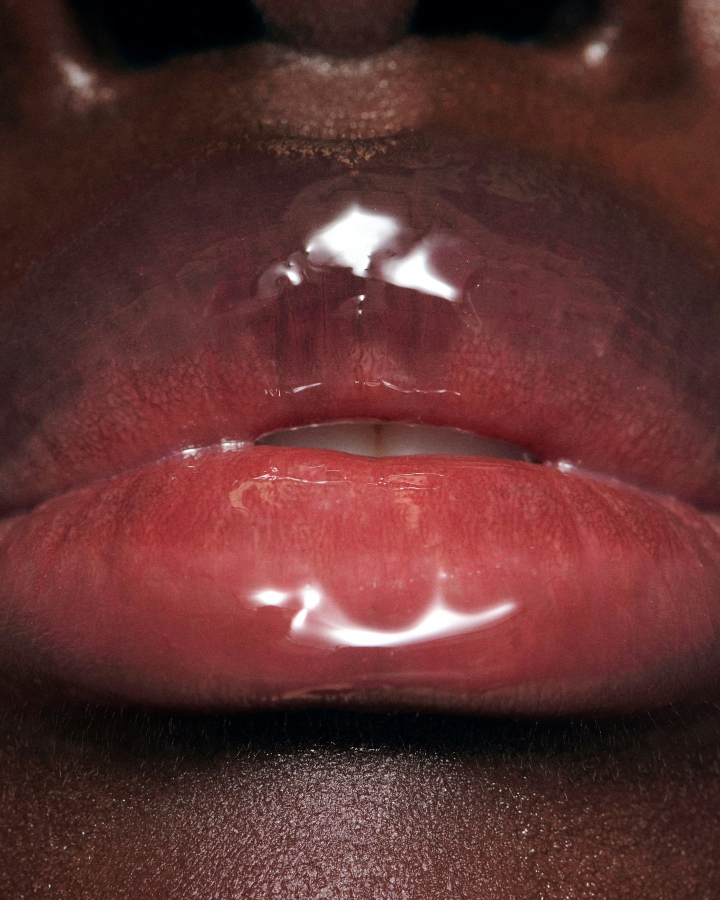 Close-up of a woman's dark skin around bright red lips with clear glossy lip gloss, showcasing texture and color.