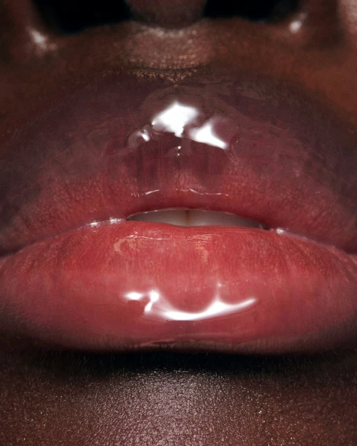 Close-up of a woman's dark skin around bright red lips with clear glossy lip gloss, showcasing texture and color.