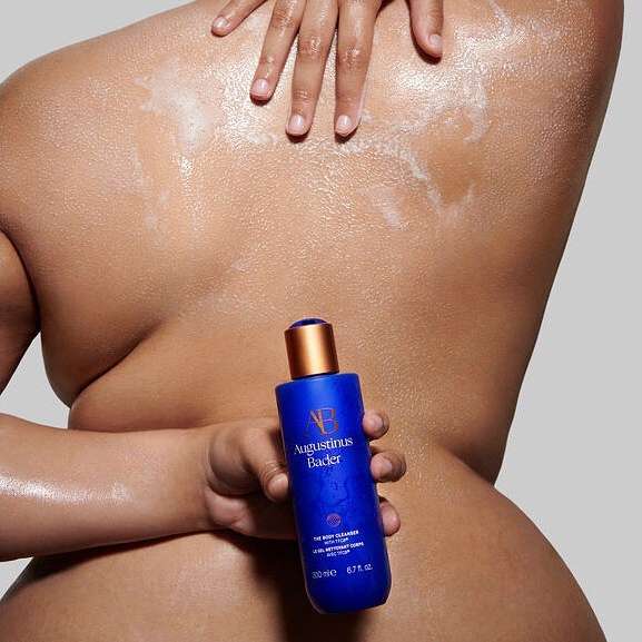 Close-up of a person applying body cleanser to their back, holding a blue bottle of Augustinus Bader body cleanser product.