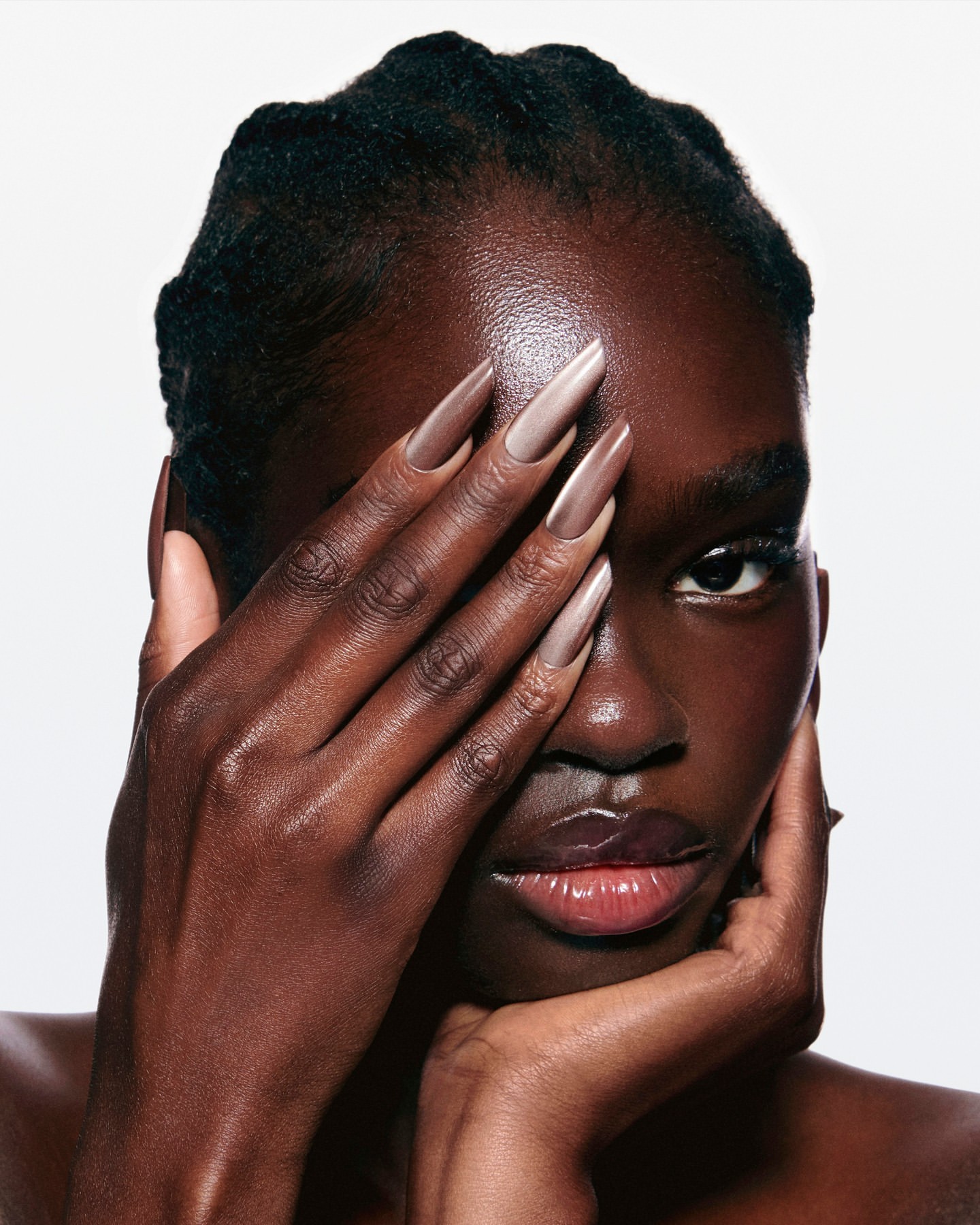 Close-up portrait of a dark-skinned woman with taupe nail polish covering one eye, fair lighting, against a plain white background.