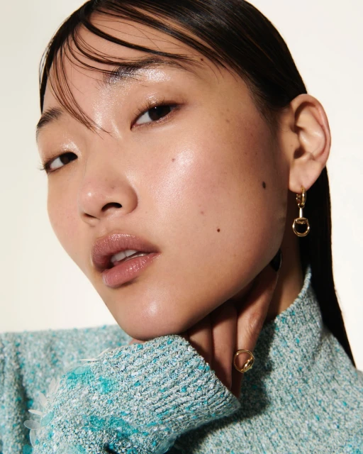 Close-up portrait of a young Asian woman wearing a textured turquoise sweater and gold jewelry in front of a white background.