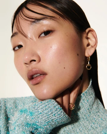 Close-up portrait of a young Asian woman wearing a textured turquoise sweater and gold jewelry in front of a white background.