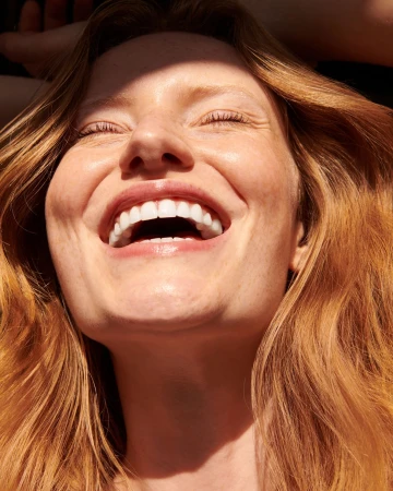 Close-up portrait of a smiling woman with red hair and freckles in bright natural sunlight.