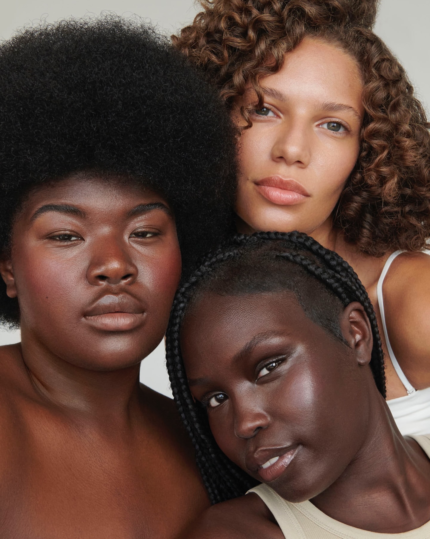 Three women with varying skin tones and hair textures pose in a close-up beauty shot against a neutral backdrop, showcasing natural makeup.