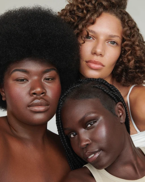 Three women with varying skin tones and hair textures pose in a close-up beauty shot against a neutral backdrop, showcasing natural makeup.