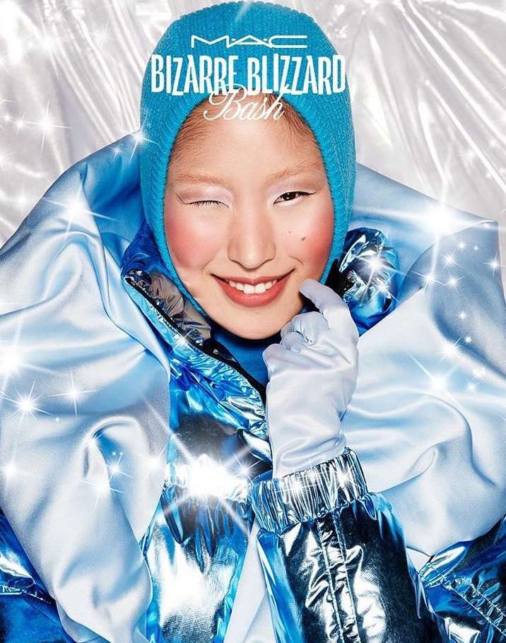 Smiling adult woman with light skin and blue winter cap winks at the camera wearing reflective blue jacket in front of silver backdrop.