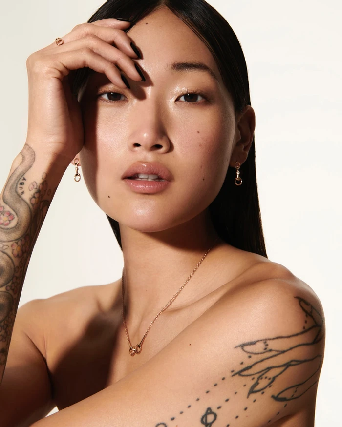 Close-up portrait of an East Asian woman with dark hair, tattoos on her arms, and gold jewelry against a white background.