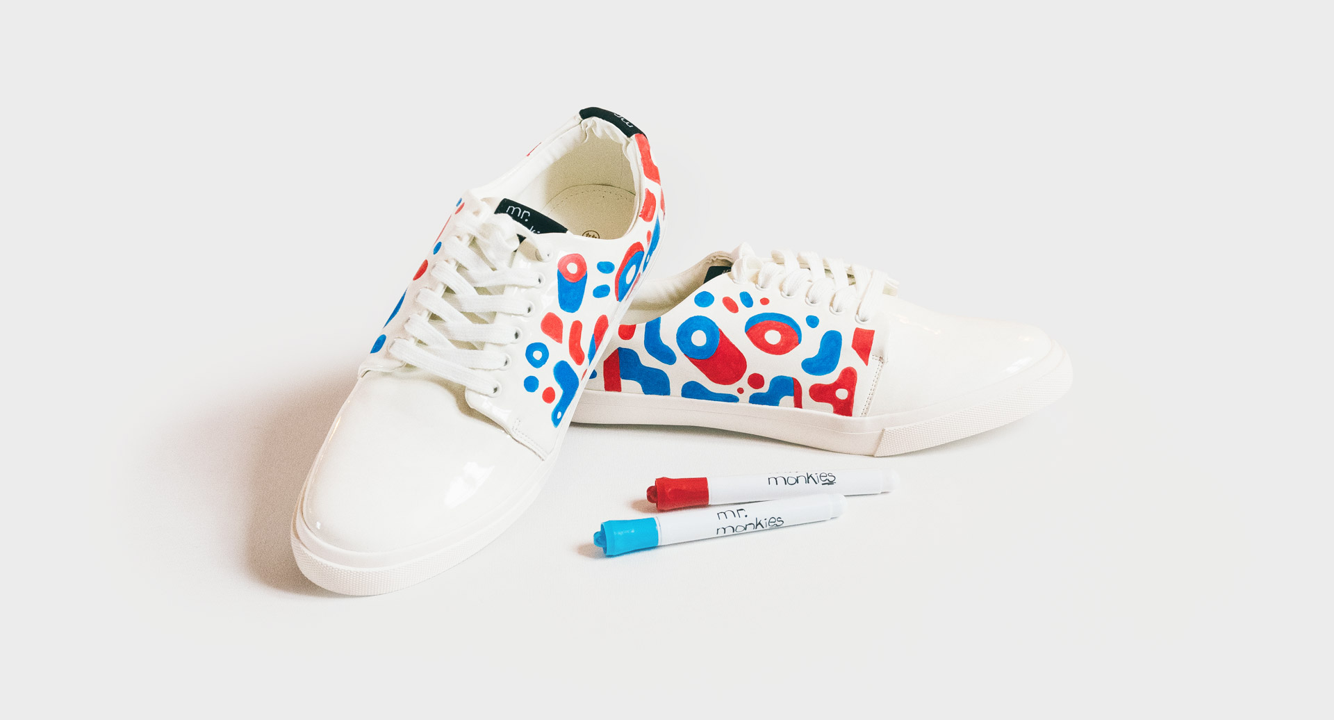 Two white sneakers are decorated with red and blue abstract designs, accompanied by two markers, presented against a white backdrop.