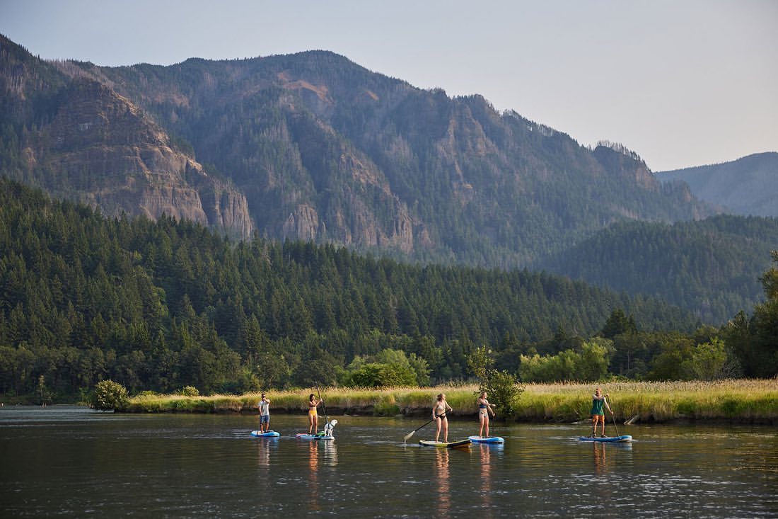 Group paddleboarding on river with mountain backdrop. People stand on boards, paddling on smooth water, enjoying outdoor recreation.