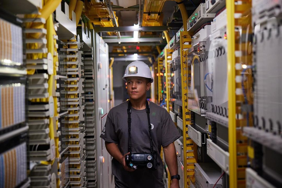 Adult in a hard hat and work clothes stands in a telecommunications room filled with rows of yellow cable racks and network servers.