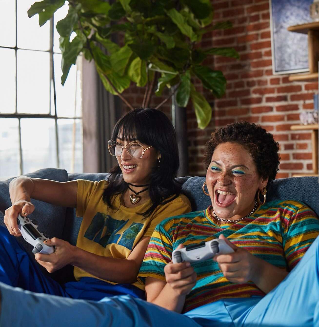 Two cheerful young women play a video game together on a blue sofa in a brightly lit living room with exposed brick and a large plant.
