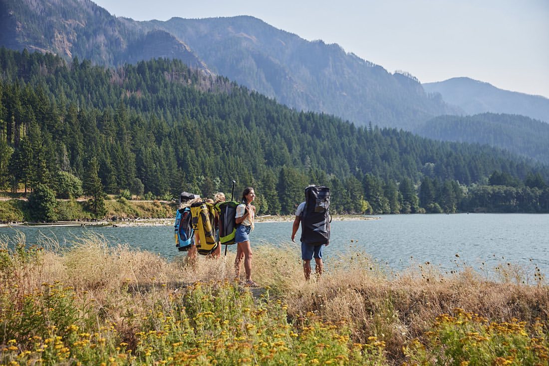Group of friends hiking with large backpacks on a trail by a lake, with mountains and trees in the background on a sunny day.
