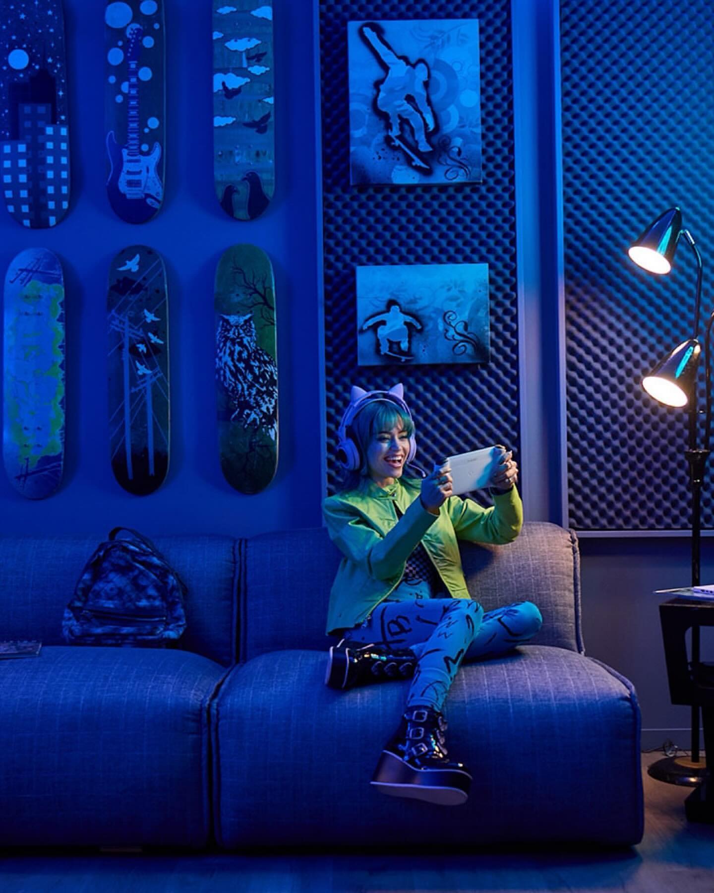Young woman playing a handheld video game while seated on a sofa in a room with skateboards and artwork on the walls. 