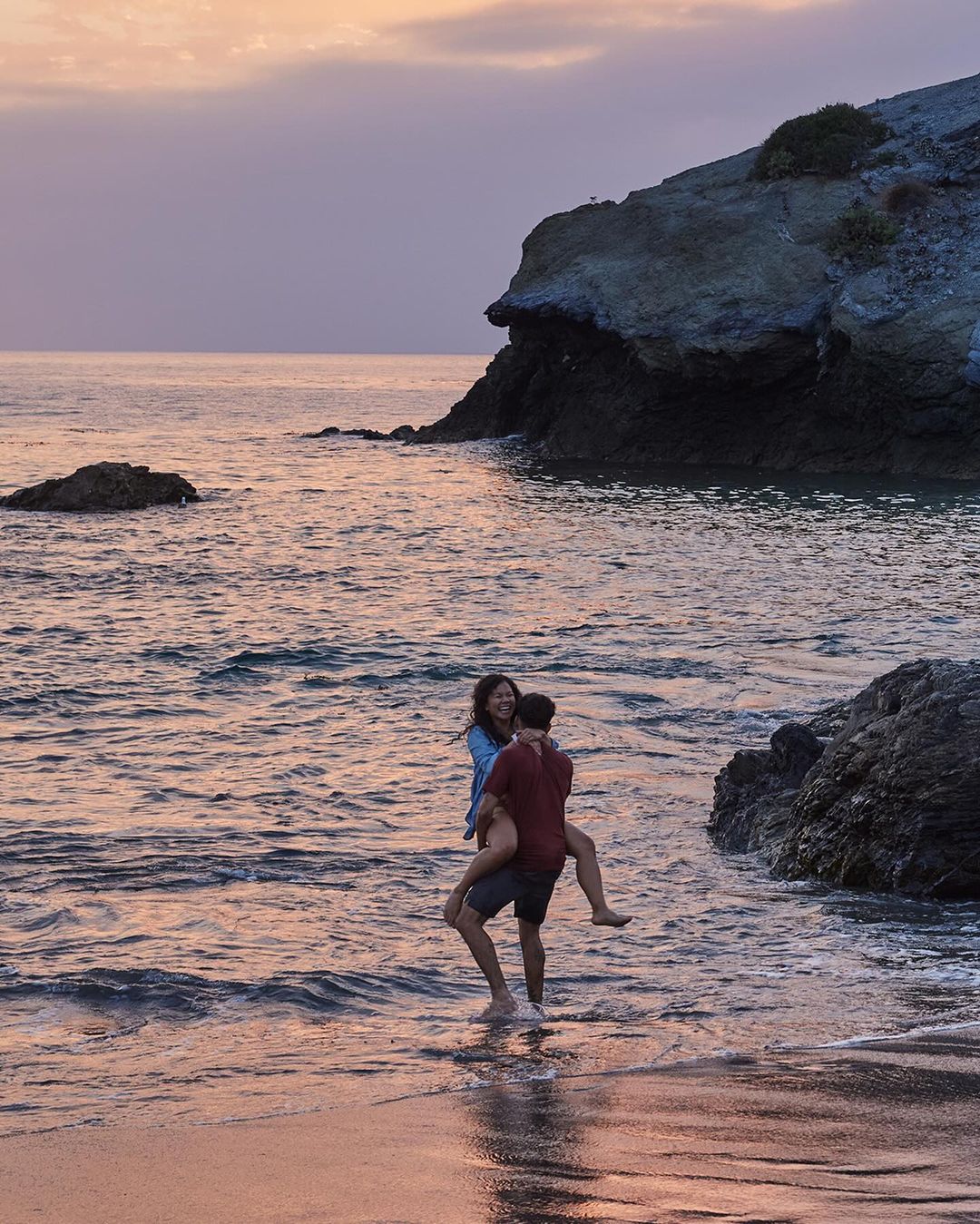 A couple enjoys time on a beach at sunset with a man giving a woman a piggyback ride in the water, with cliffs in the background.