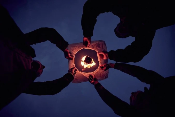 Five people release a paper lantern into the night sky, creating a warm glow against the dark blue backdrop.
