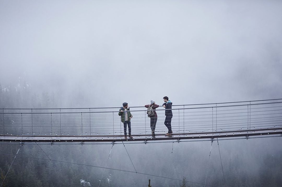 Three people stand on a suspension bridge surrounded by a forest and fog, creating a scenic and misty outdoor environment.