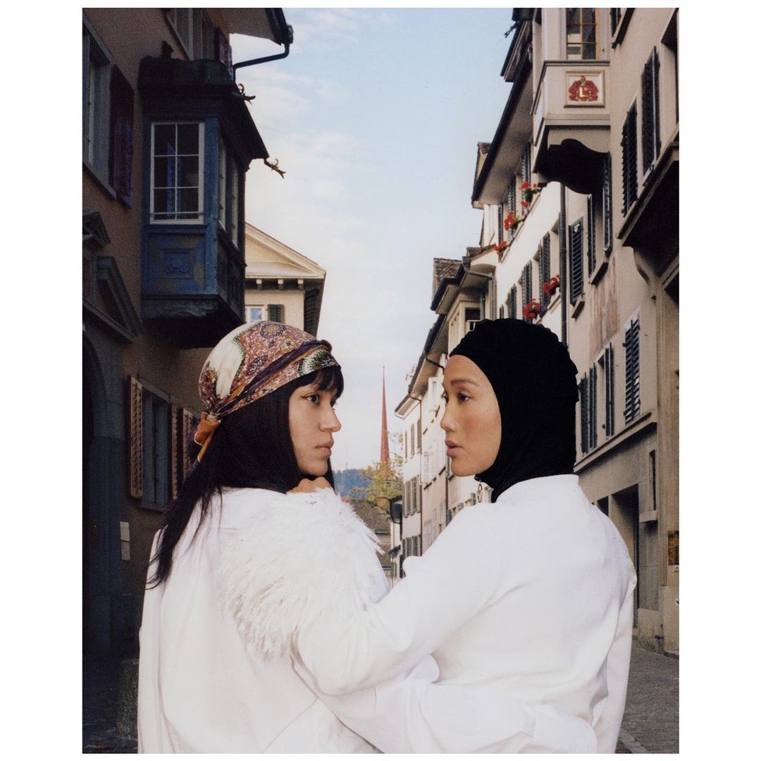 Two Asian women in white coats pose in a European city. One wears a headscarf, the other wears a bandana. Urban fashion and architecture.