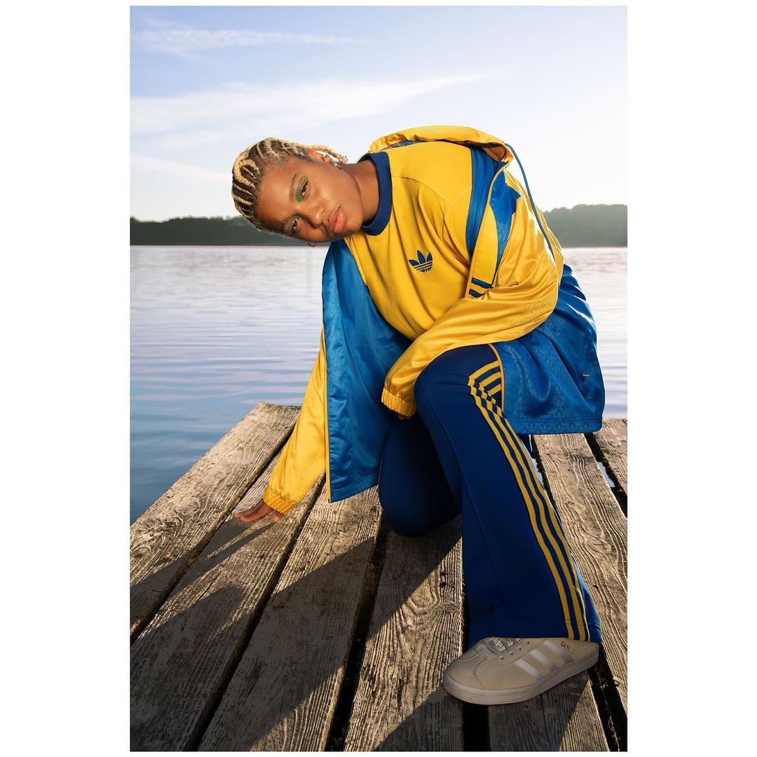 Woman poses on dock wearing blue and yellow Adidas track suit against lake and sky background in an eye-level medium shot.