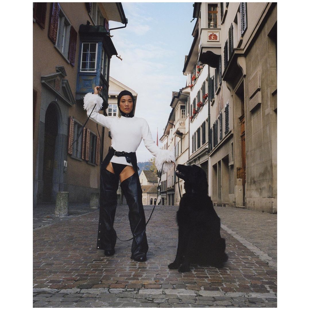 Woman and black dog pose on European brick street wearing chaps and white sweater with long sleeves with fur trim cuffs.