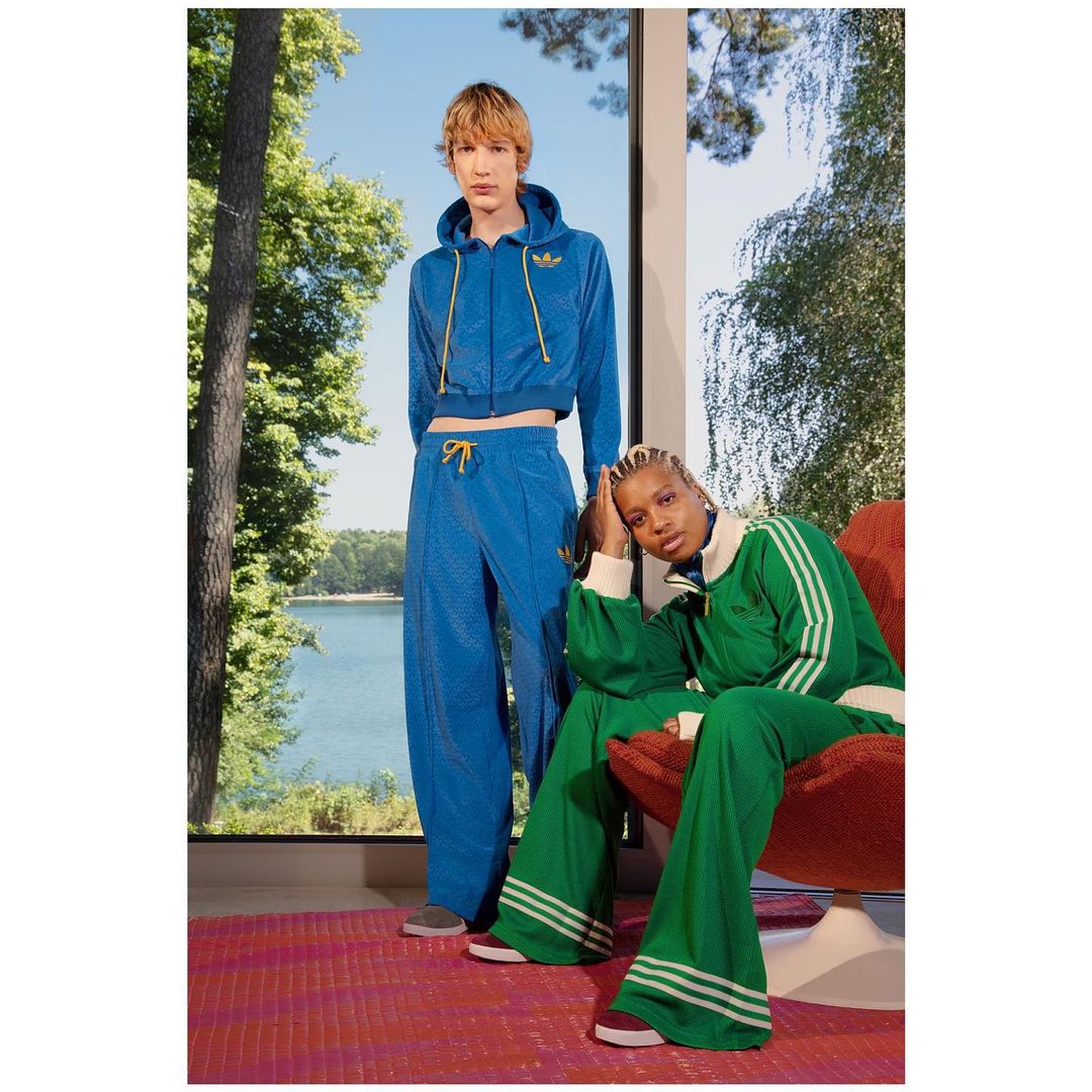 Two people in matching athletic tracksuits pose indoors near a large window with a nature view. A person is seated, and the other stands.