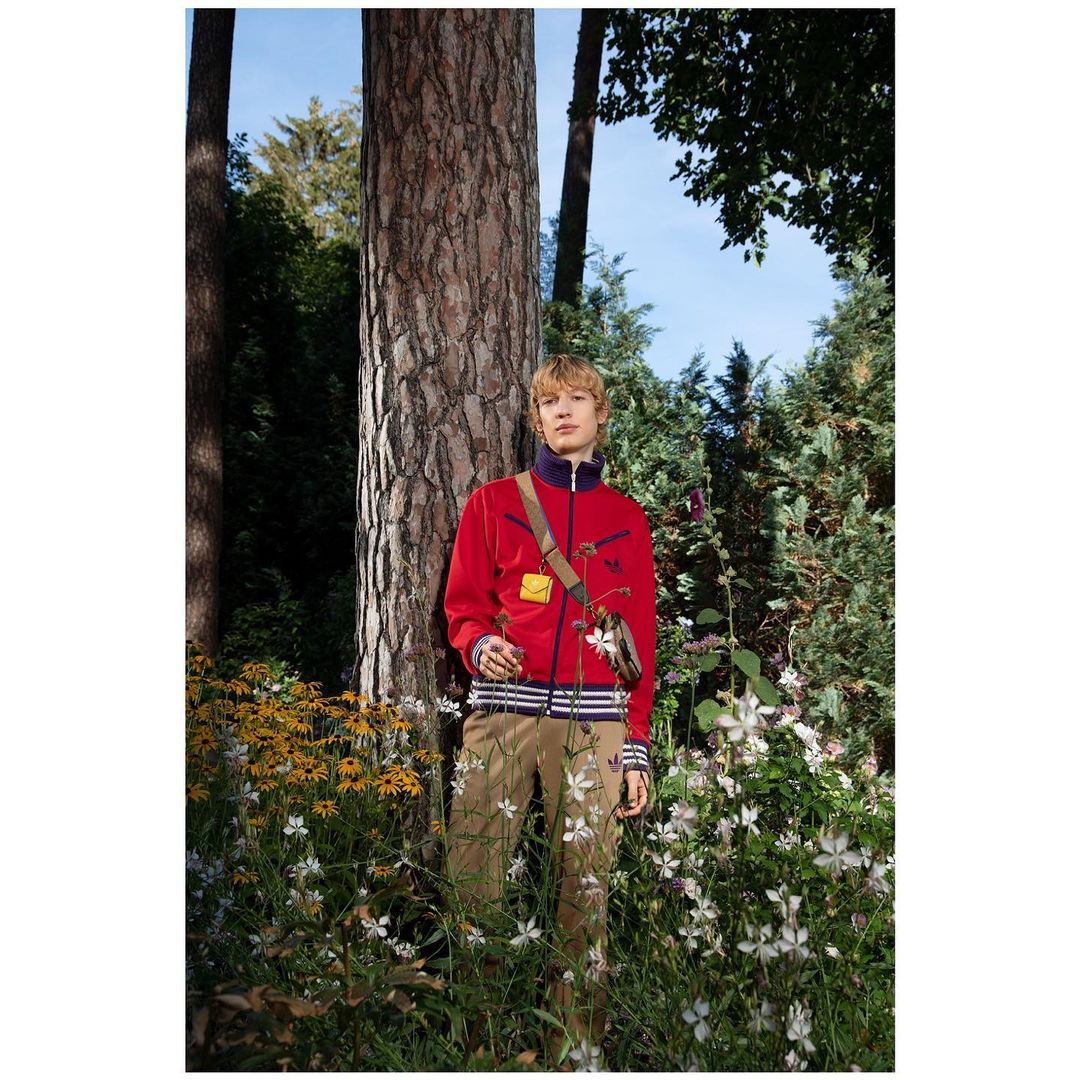 Man in red tracksuit standing next to a tree in a garden with white and yellow flowers, wearing a small shoulder bag and tan pants.