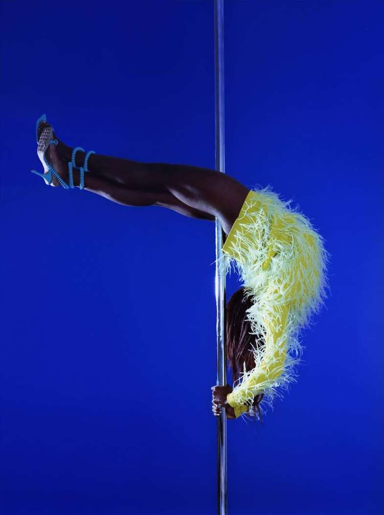 A slender woman showcases her fitness and flexibility on a pole while wearing a feathered yellow outfit against a blue background.