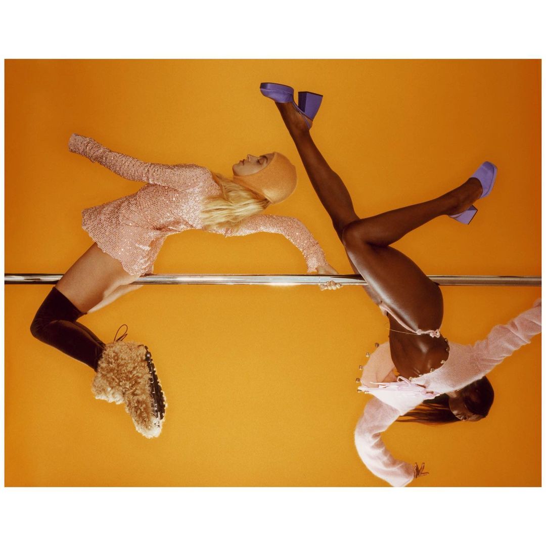 Two fashion models perform choreography in a studio with an orange backdrop, emphasizing their stylish outfits and dynamic poses.